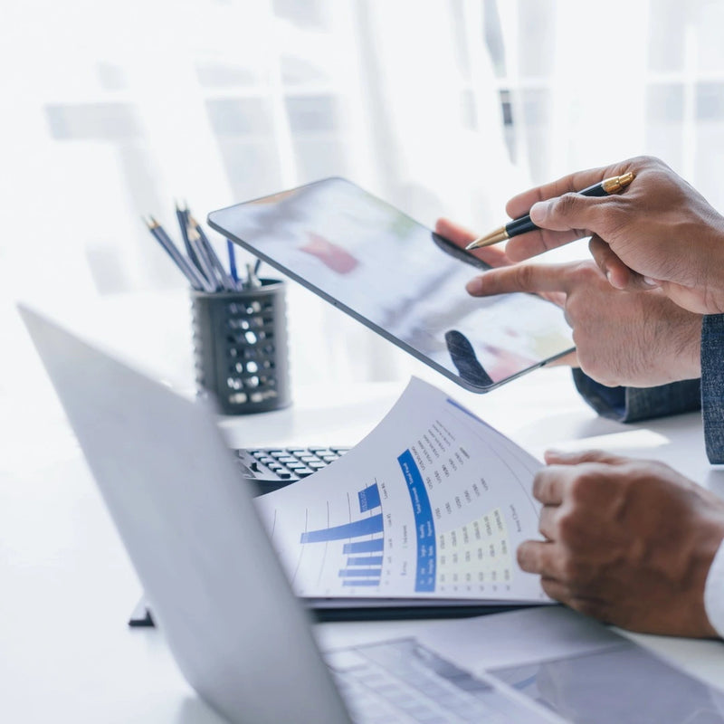 Person holding a tablet and looking at printed documents with a laptop on a desk.